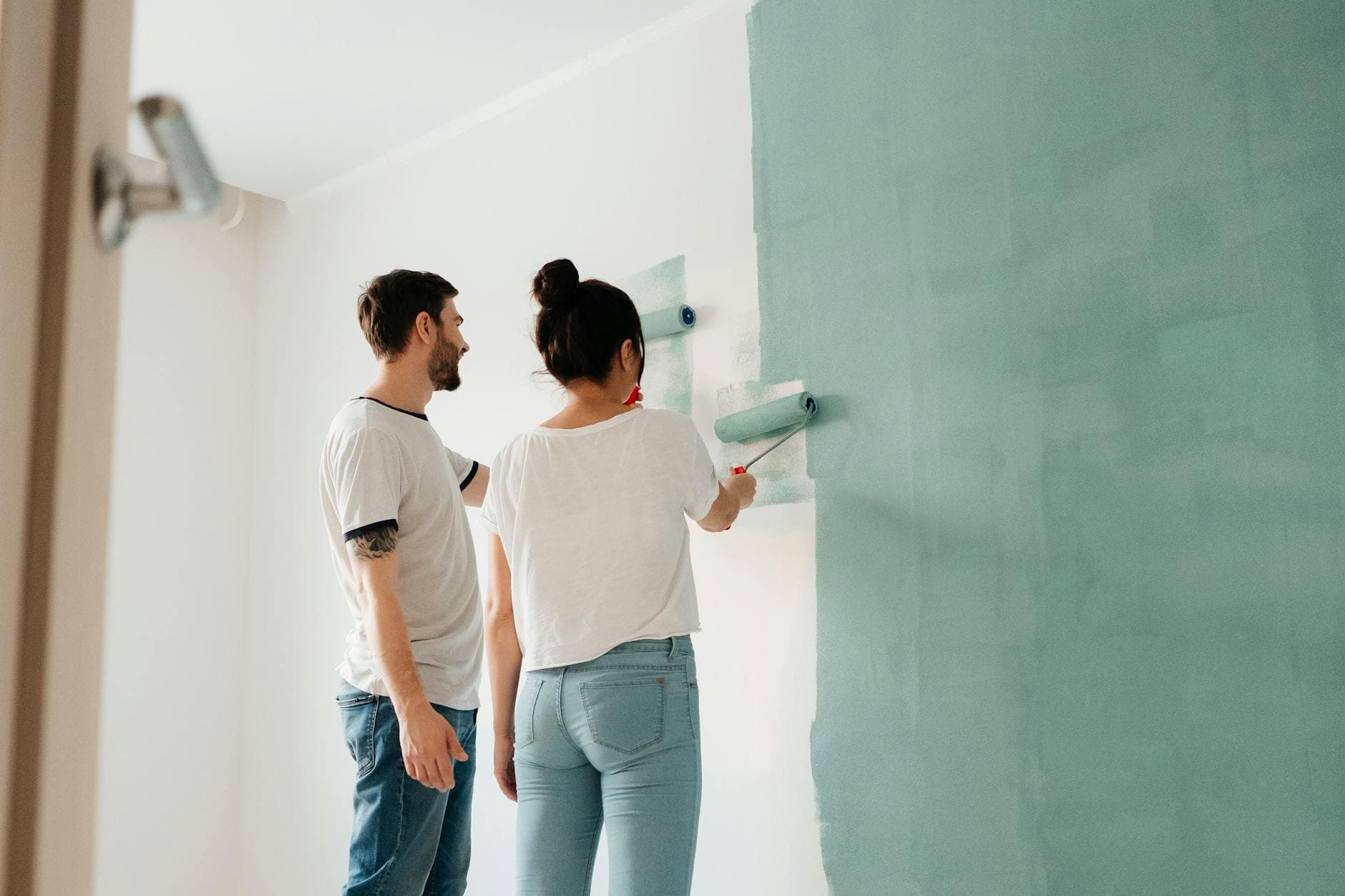 A couple collaborates to paint a wall with a roller indoors, showcasing teamwork.