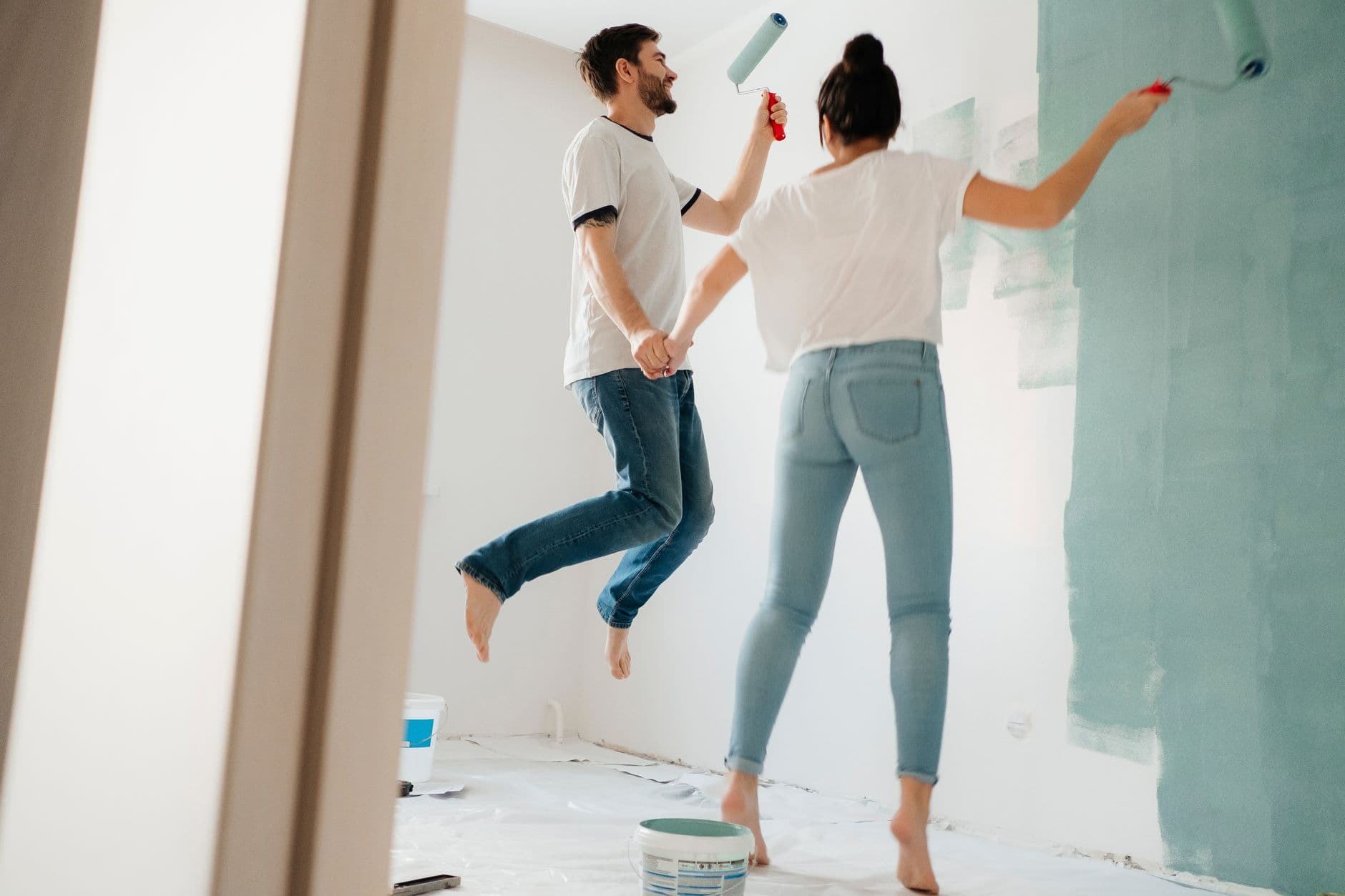 A playful couple painting walls together in their home, having fun while holding hands.
